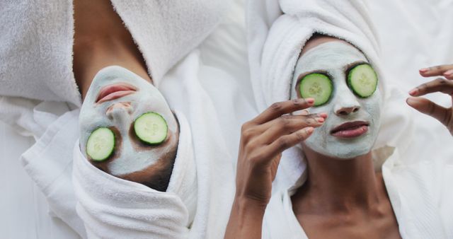 Image of relaxed diverse female friends moisturizing with face masks and cucumbers. Friendship and taking care of yourself and beauty concept.
