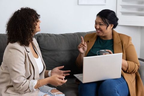 Diverse female colleagues discussing project on gray sofa with laptop and coffee