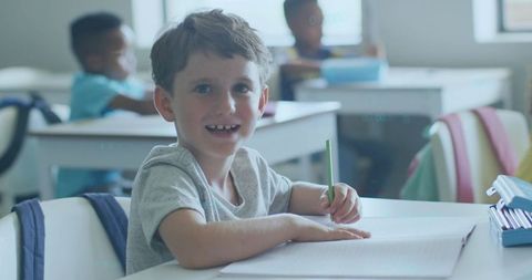 Smiling elementary student writing with green pencil at desk, backpacks in background