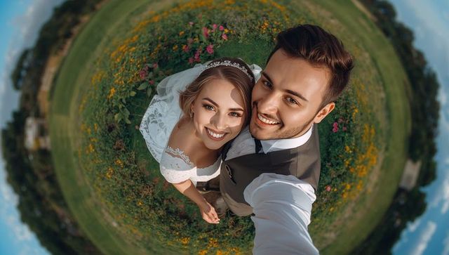 Smiling newlyweds taking fisheye selfie in wildflower meadow showing bridal dress veil
