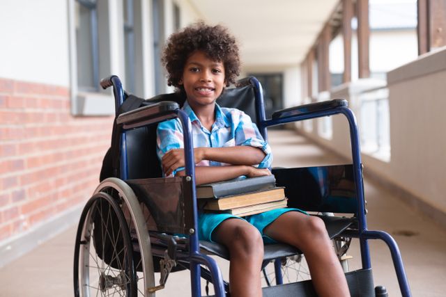 Young African American boy sitting in a wheelchair, holding books and smiling in a school corridor. Ideal for use in educational materials, inclusive education campaigns, disability awareness, and back-to-school promotions.