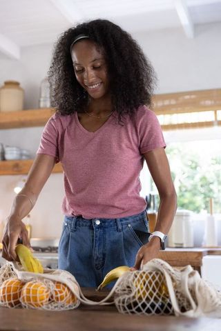 African american woman packing bananas and oranges into reusable mesh bags on kitchen island