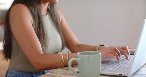 Young woman typing on laptop at home desk with green mug, gold bangles and wristwatch