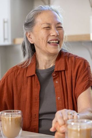 Senior asian woman laughing at kitchen table with coffee, warm candid lifestyle moment