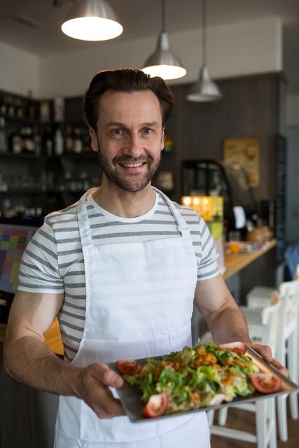 Waiter carrying a tray of fresh salad in a restaurant, smiling at the camera. Ideal for use in articles or advertisements related to dining, hospitality, healthy eating, restaurant services, and culinary arts.