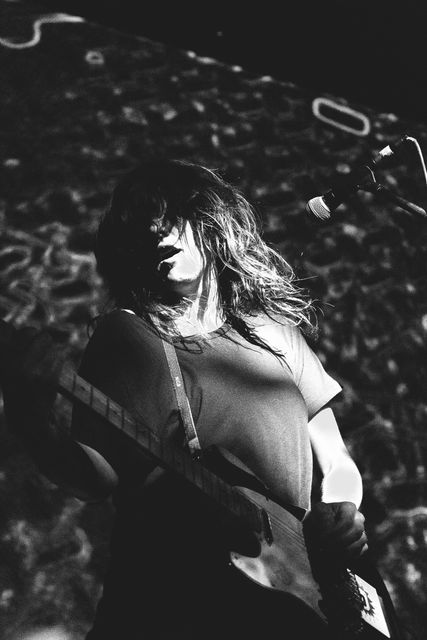 Woman rock musician playing electric guitar and singing into microphone on stage. Black and white photo captures intense performance energy and passion. Suitable for use in music event promotions, concert posters, musician profiles, and articles about live music performances.