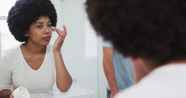 Woman standing in front of a bathroom mirror applying moisturizer to her face. Reflective surface showing her focusing on her skincare routine. Ideal for use in skincare product advertisements, beauty and self-care promotional materials, or articles about daily grooming routines and personal hygiene tips.