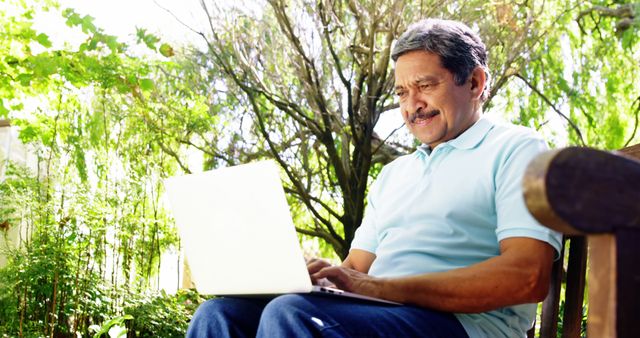 Older man sitting outside on bench typing on laptop. Green surroundings with sunlight filtering through trees. Useful for topics on seniors and technology, enjoying nature, outdoor productivity, remote working, retirement activities, and active senior lifestyles.