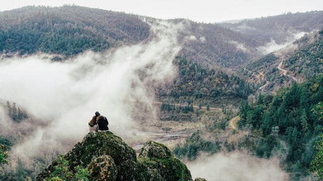 Couple sitting on rocky outcrop overlooking misty valley, surrounded by forested mountains. Ideal for travel guides, nature retreats, adventure blogging, or outdoor lifestyle promotions.