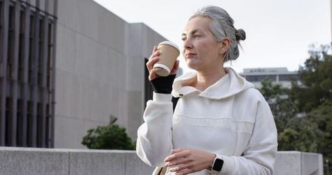 Mature woman sipping takeaway coffee on urban terrace wearing hoodie and smartwatch