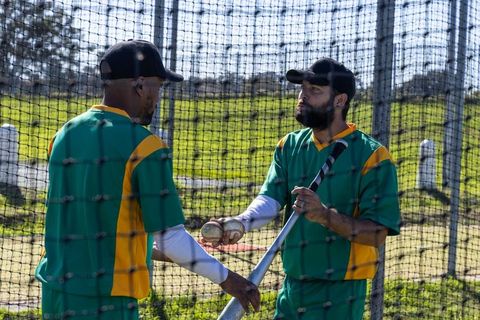 Teammates practicing batting drill in outdoor cage passing baseballs and holding bat