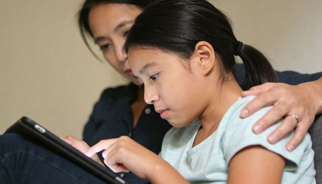 Mother Guiding Daughter Using Tablet, Daughter Focusing and Tapping Screen on Couch