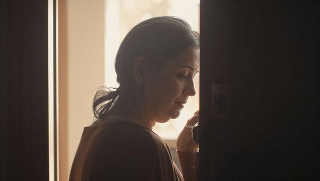 Pensive woman standing at doorway holding doorknob backlit by warm window light