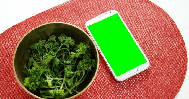 Mockup with smartphone with green screen next to fresh parsley in bowl. Useful for creating food-related content, technology marketing, organic lifestyle presentations, healthy eating promotions.