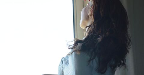 Backlit woman standing by bright window facing sunlight with long hair and blue blouse