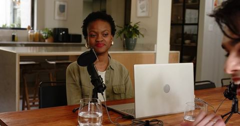 Two hosts recording cozy home podcast in kitchen with laptop, microphones, natural light