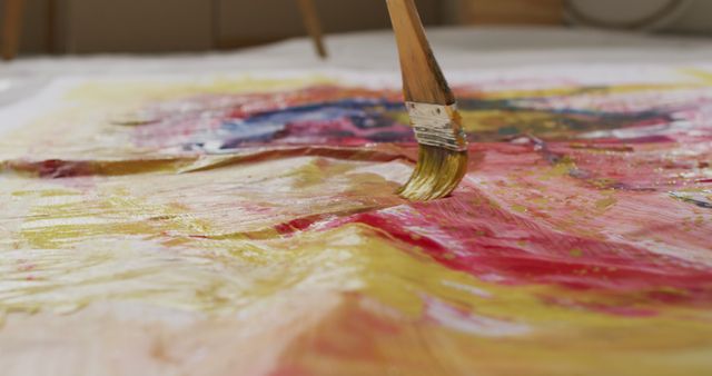 Close-up of artist using large brush to paint abstract work on canvas featuring bright colors like yellow, red, and blue. Perfect for illustrating concepts of creativity, contemporary art, and the artistic process. Ideal for use in art tutorials, creative design projects, or advertising for painting workshops.