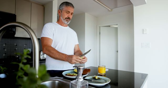Mature man is peeling fruit over the counter in a modern, bright kitchen. The countertop includes a plate with food, a glass of juice, and a hand soap dispenser. Man is engaged, showing concentration while preparing a healthy breakfast, showcasing a healthy lifestyle. Ideal for use in campaigns centered around healthy living, home life, morning routines, and food preparation.