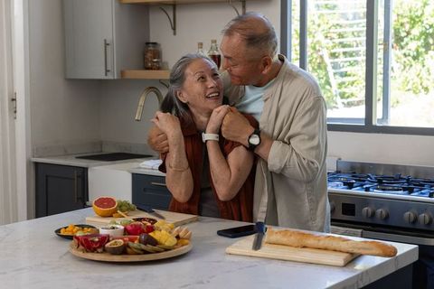 Sunlit senior asian couple embracing at marble island with fruit platter and baguette