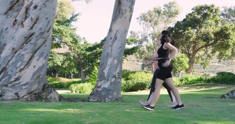 Two women power-walking through sunlit park wearing activewear and carrying water bottle
