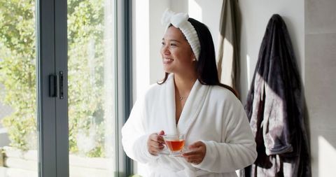 Woman in robe enjoying tea relaxing by sunlit window