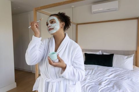 African american woman applying mint face mask in bedroom wearing white bathrobe