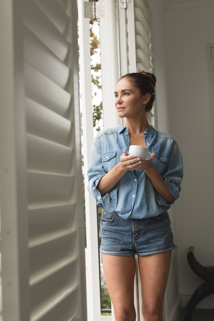 This image depicts a woman standing by a window, holding a cup of coffee, and enjoying a peaceful moment in a cozy home. She is dressed casually in a denim shirt and shorts, suggesting a relaxed and comfortable atmosphere. This image can be used for lifestyle blogs, home decor websites, or advertisements promoting relaxation and comfort.
