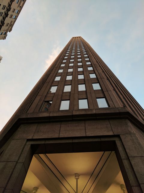 Upward view captures the sheer height and architectural design of a modern office building against a sunset sky. Suitable for themes related to urban development, business environments, corporate offices, and modern architecture. Ideal for advertisements, websites, and articles focusing on city landscapes and high-rise office spaces.