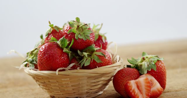 A basket filled with ripe strawberries sits on a wooden surface, with one strawberry cut in half beside it. Fresh strawberries like these are often used in culinary dishes for their sweet flavor and vibrant color.