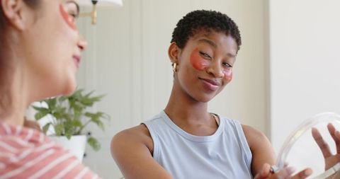 African american woman applying red hydrogel eye patches while holding mirror, smiling