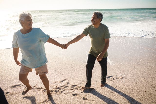 Senior couple holding hands while walking on the beach. They are smiling and enjoying their time together by the ocean. This image can be used for promoting senior lifestyle, retirement plans, travel destinations, and health and wellness for older adults.