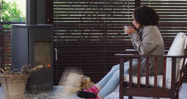 Woman is sitting in a wooden chair by the fireplace, enjoying a hot beverage while surrounded by cozy and warm indoor atmosphere. This scene is ideal for advertisements related to comfort, winter clothing, hot drinks, cozy home settings, and indoor relaxation.