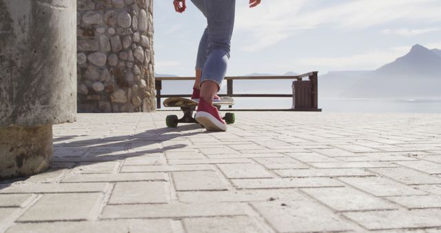 Legs of person wearing jeans and red sneakers skateboarding near coastal area on sunny day. This can be used for content relating to outdoor activities, leisure pursuits, or skateboarding enthusiasts. It invokes themes of adventure, youth, and freedom, making it ideal for travel blogs, lifestyle brands, or sports promotions.