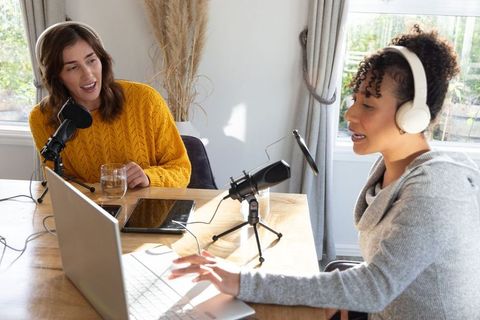 Women podcasters recording casual home podcast wearing headphones and sweaters with laptop
