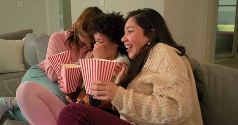 Multiracial female family laughing and holding popcorn during cozy movie night on sofa