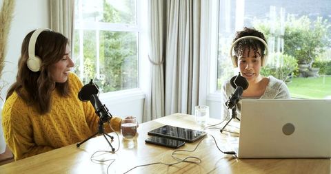 Diverse female co-hosts recording podcast at home with natural light, microphones, laptop