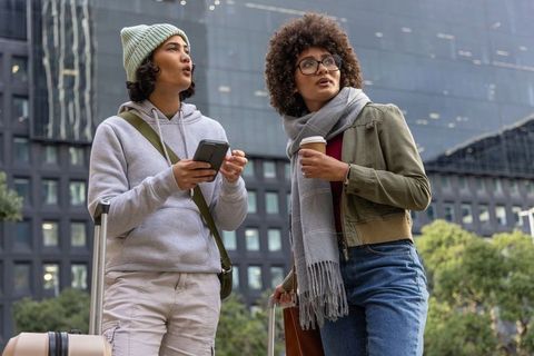 Young women traveling with luggage in urban downtown plaza using smartphone and coffee