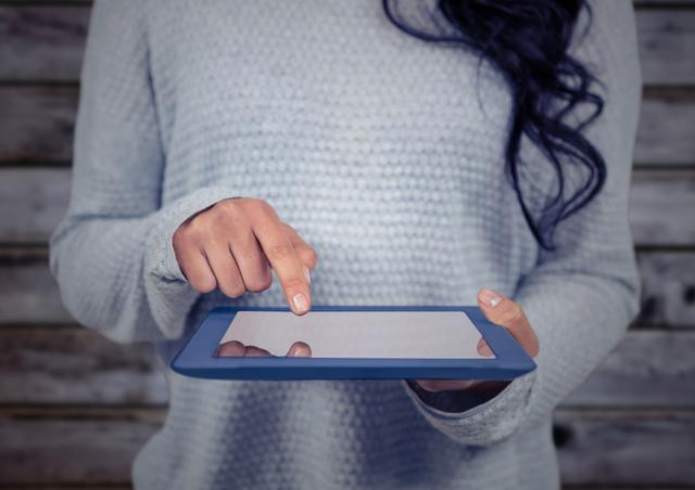 Woman holding and using digital tablet while wearing casual gray sweater against wooden background. Ideal for technology, digital lifestyle, online learning, and casual daily use themes.