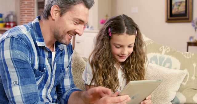 Father and daughter enjoying time together on a tablet in a cozy living room. Both smiling and interacting with the device. Ideal for advertising family services, technology products, parenting blogs, and family-oriented content.