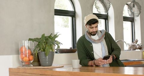Indian man leaning on wooden counter checking smartphone in cozy sunlit kitchen