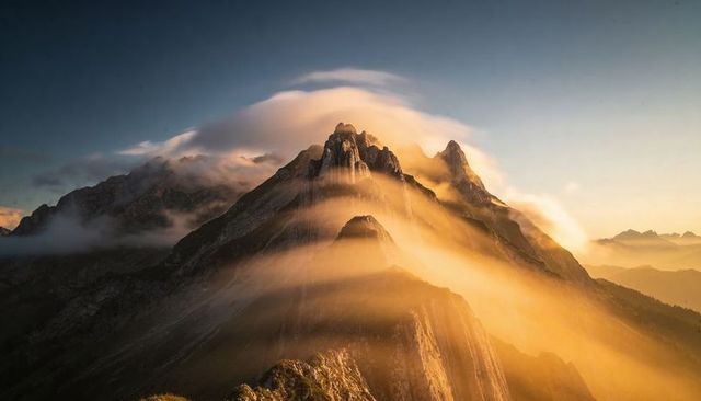 Golden light glowing over jagged alpine peak with lenticular cloud cap at sunrise