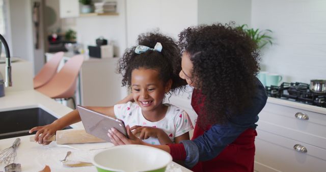Happy scene of mother and daughter baking together, watching a tablet in a modern kitchen. Ideal for family-related content, online cooking tutorials, or home appliance advertisements. Perfect image to showcase family bonding, educational activities, and modern home life.