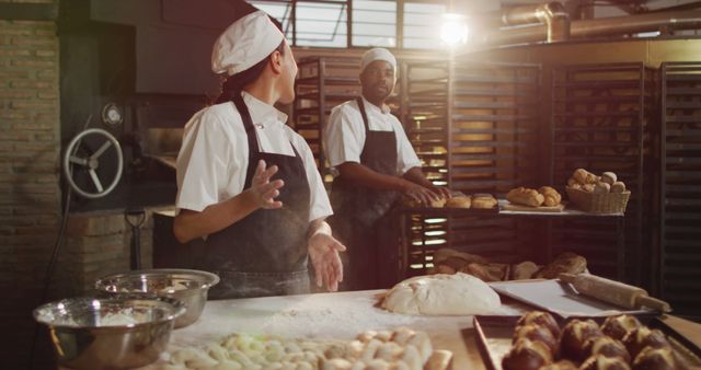 Two bakers in professional attire discussing work in an artisan bakery. The setting shows a countertop covered in flour and dough, with various baked goods and pastries around. Useful for content related to teamwork, culinary arts, and professional baking.