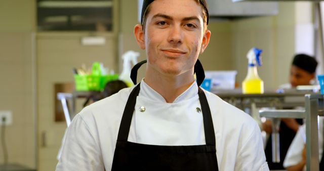 Young male chef standing confidently in a professional kitchen environment, dressed in a chef's uniform and apron. Ideal for use in articles or advertisements related to culinary education, restaurant businesses, hospitality industry, and cooking tutorials.