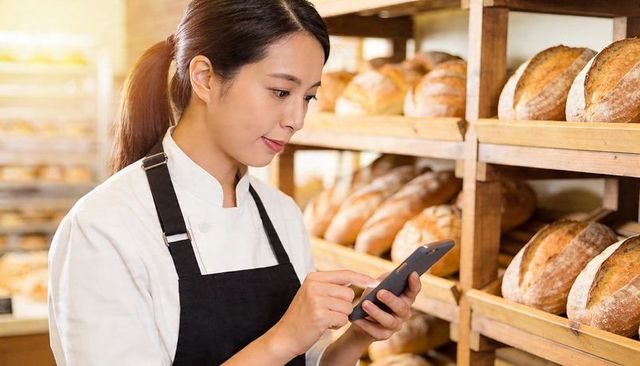 Baker Checking Smartphone While Managing Orders in Artisanal Bakery with Wooden Racks