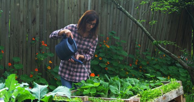 Woman watering plants in a backyard garden, surrounded by greenery and wooden fence. Ideal for themes related to home gardening, outdoor activities, nature, self-sufficiency, and healthy living.