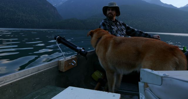 A Caucasian man in a cowboy hat enjoys a boat ride with his dog amidst a scenic mountain lake, with copy space. His relaxed posture and the serene environment suggest a peaceful fishing trip or leisurely outing.