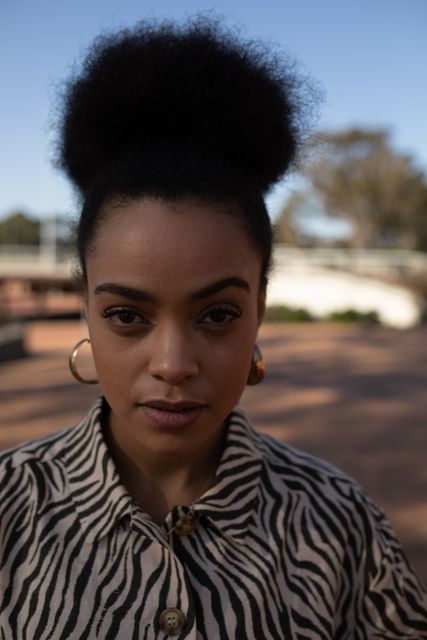 This image captures a confident biracial woman enjoying a sunny day in the city. She is wearing a zebra print shirt and gold earrings, with her natural hair styled in an afro. This photo can be used for fashion editorials, lifestyle blogs, urban living promotions, or advertisements focusing on diversity and confidence.