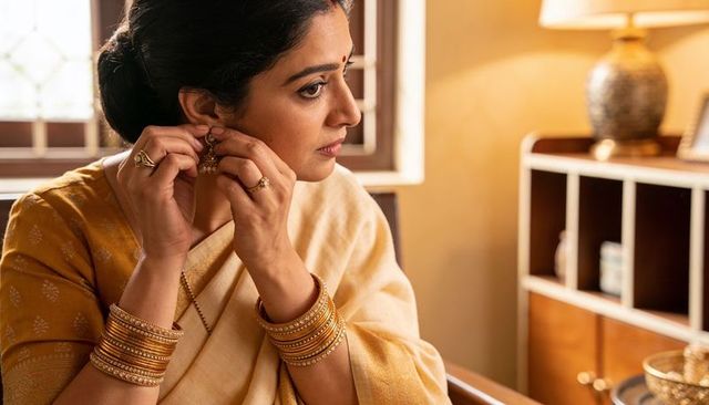 Indian woman adjusting gold earring wearing cream sari and stacked gold bangles by window