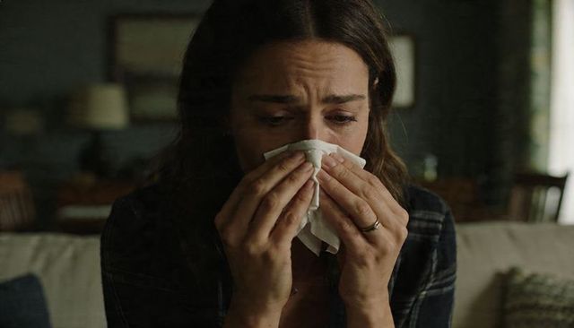 Tearful woman sitting on couch wiping nose with tissue, wedding ring visible